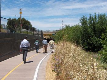 A person in business clothes walks slightly behind 4 other people in business clothes away from the camera down a paved walking trail with painted white and yellow divider lines, framed with grass and vegetation on the right side and a concrete wall on the left side