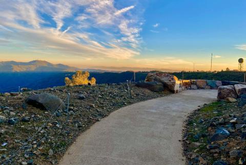 A paved trail curves around a rocky hilltop with a magnificent view of blue mountains all around, under a bright blue sky with a hint of yellow on the horizon