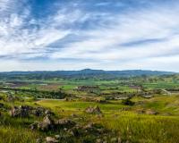 Looking over green hills across a valley below filled with agricultural fields, to blue mountains in the distance, under a blue sky streaked with white clouds 