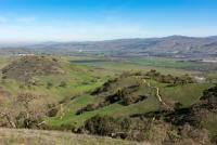 Rolling green hills with oak trees and winding trails overlooking a wide green valley beyond them, and mountains on the far side, under a blue sky.