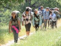 A line of girls wearing backpacking backpacks walk along a trail bordered by tall grass, smiling at the camera