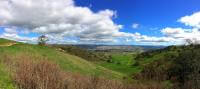 A panoramic photo of green hillsides with hiking trails overlooking a wide green valley, with mountains in the distance, under a blue sky with white fluffy clouds